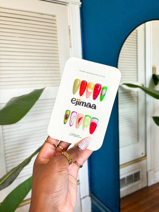 Hand holding a card with strawberry, lemon, passionfruit, lemon, avocado, red chili, watermelon and lime designs. Press on nails, custom nail set with 'ejimaa' branding in front of a mirror.