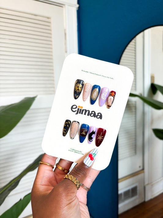 Hand holding a nail card with a charmed tv series inspired themed set for halloween and 'ejimaa' branding against a home interior background.