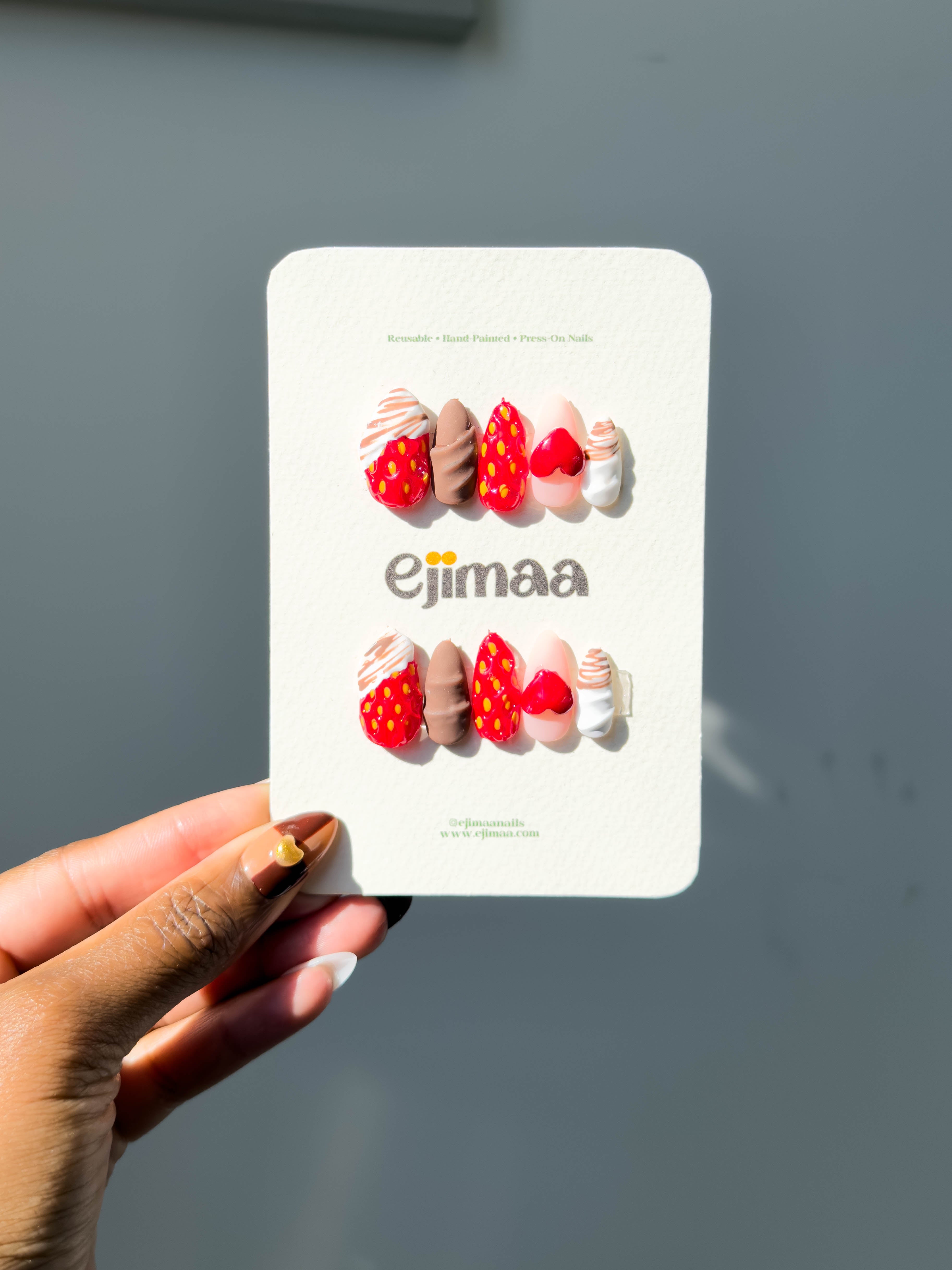 Hand holding a card with strawberry-themed nails and 'ejimaa' branding against a gray background
