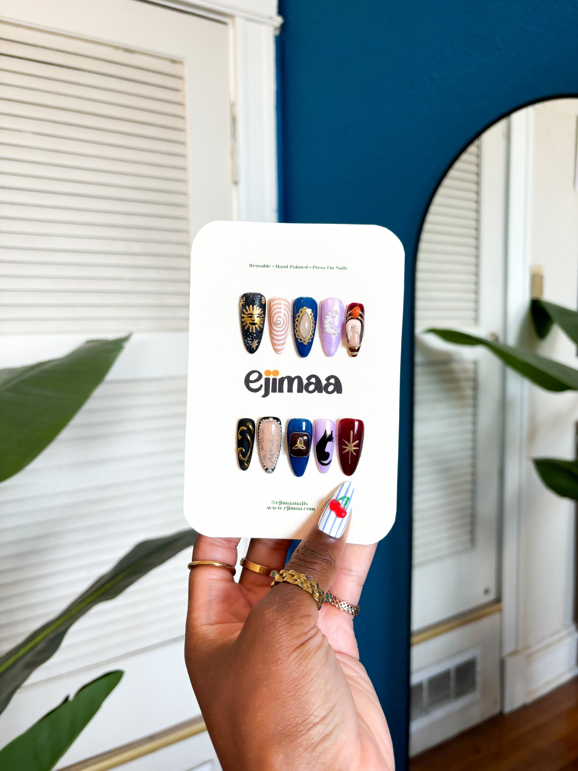 Hand holding a nail card with a charmed tv series inspired themed set for halloween and 'ejimaa' branding against a home interior background.