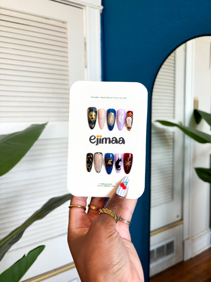 Hand holding a nail card with a charmed tv series inspired themed set for halloween and 'ejimaa' branding against a home interior background.