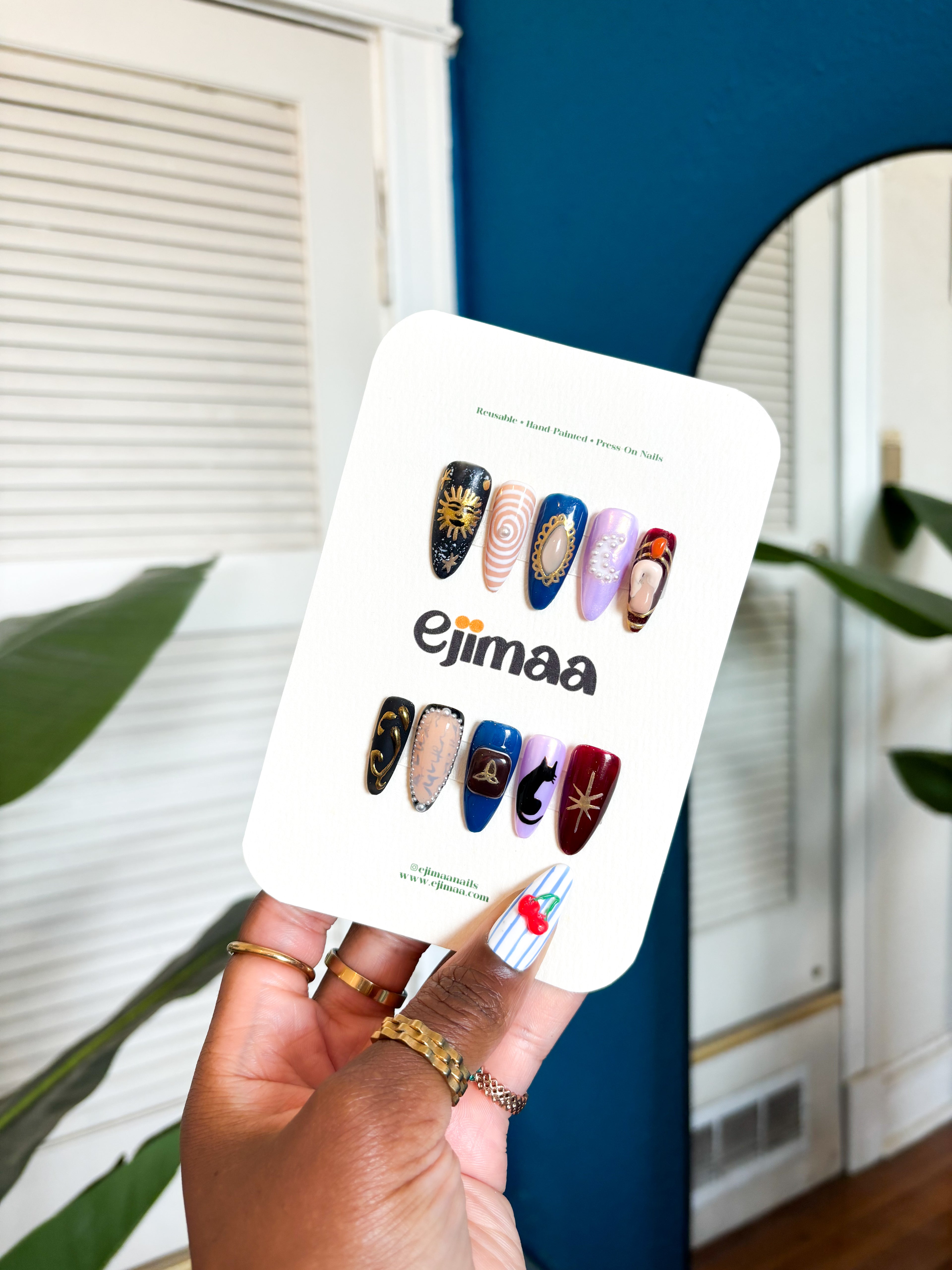 Hand holding a nail card with a charmed tv series inspired themed set for halloween and 'ejimaa' branding against a home interior background.
