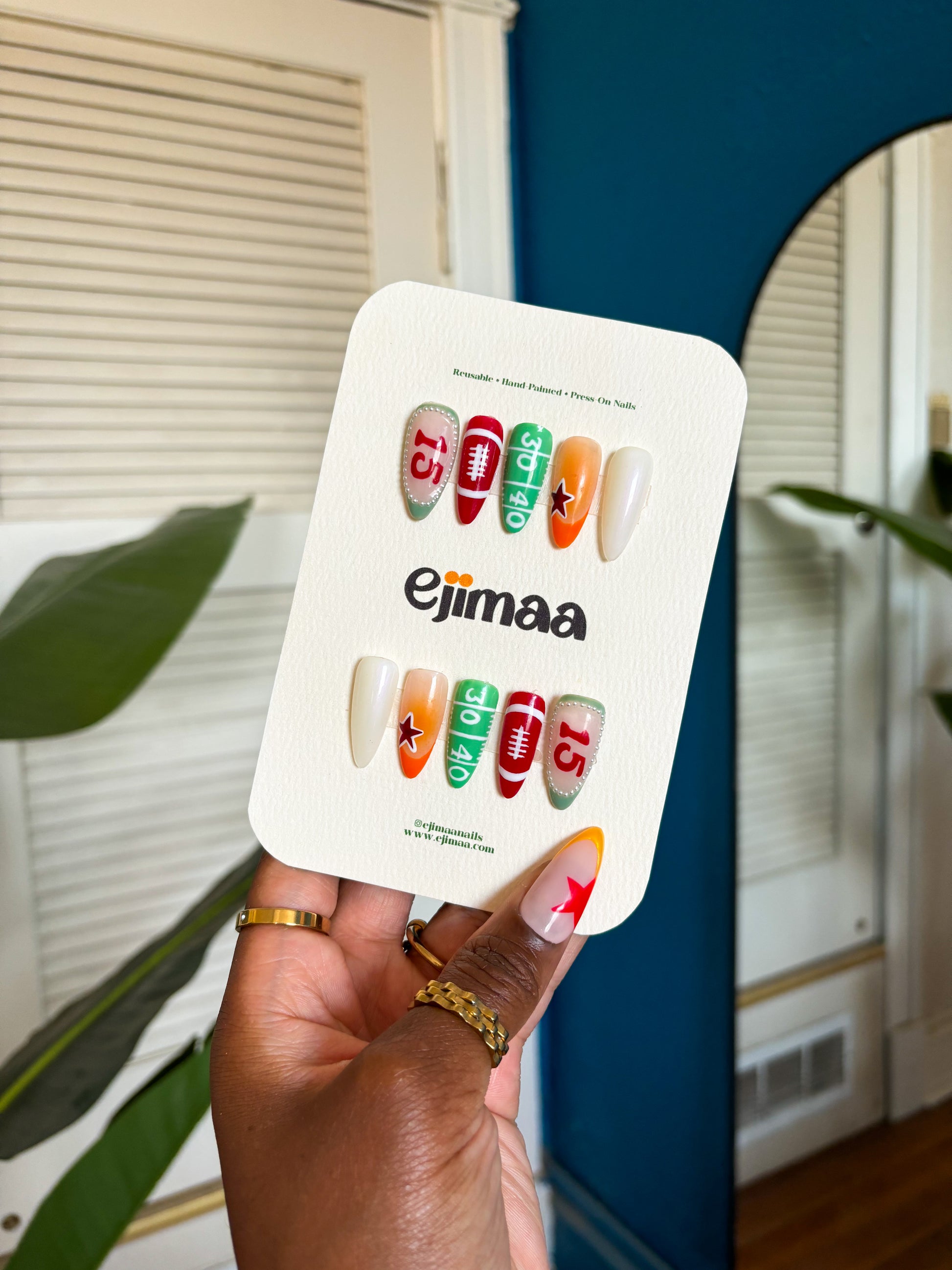 Hand holding Football Season 2025 nails. This design features a star, the number 15, pearl elements, a football and the football field. Nails are displayed on a ejimaa nail card. 