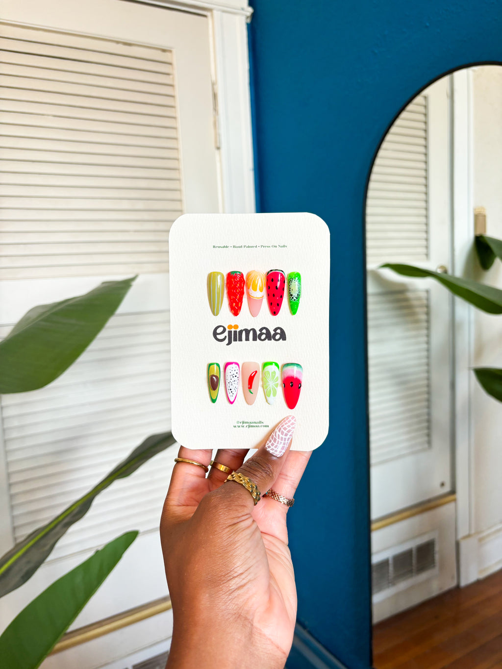 Hand holding a card with strawberry, lemon, passionfruit, lemon, avocado, red chili, watermelon and lime designs. Press on nails, custom nail set with 'ejimaa' branding in front of a mirror.