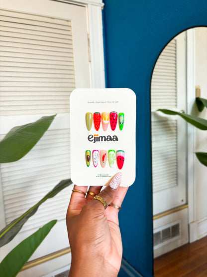 Hand holding a card with strawberry, lemon, passionfruit, lemon, avocado, red chili, watermelon and lime designs. Press on nails, custom nail set with 'ejimaa' branding in front of a mirror.